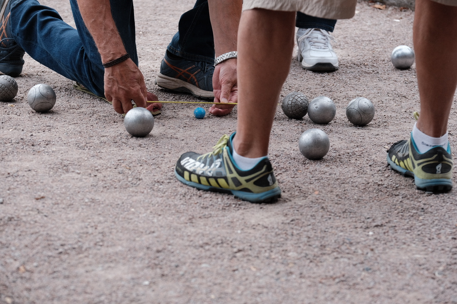 A close-up of two men crouching to measure the distance between boules on a pétanque piste with a tape measure. Only the men's feet and arms are in the image.